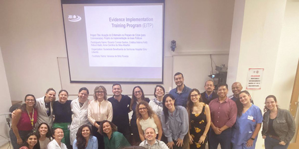 A group of photo of staff from NUEBE and JBI Brazil stand smiling in front of a presentation screen